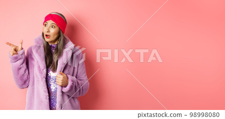 Fashion and shopping concept. Trendy elderly asian female in fake fur and glittering dress pointing, looking left with amazement, checking out offer, pink background Fashion and shopping concept. Trendy elderly asian female in fake fur and glittering dress pointing, looking left with amazement, checking out offer, pink background 98998080