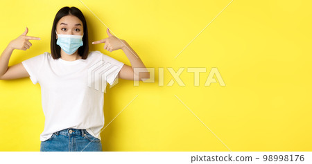 Covid-19, quarantine and social distancing concept. Young asian woman in white t-shirt, pointing at her medical mask from coronavirus, yellow background Covid-19, quarantine and social distancing concept. Young asian woman in white t-shirt, pointing at her medical mask from coronavirus, yellow background 98998176