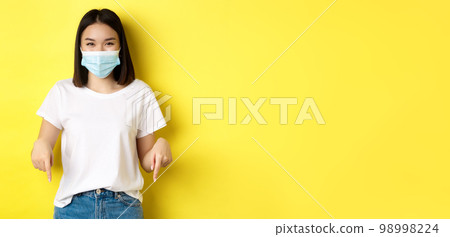 Covid-19, pandemic and social distancing concept. Happy asian woman in medical mask, showing advertisement, pointing right and smiling, yellow background Covid-19, pandemic and social distancing concept. Happy asian woman in medical mask, showing advertisement, pointing right and smiling, yellow background 98998224