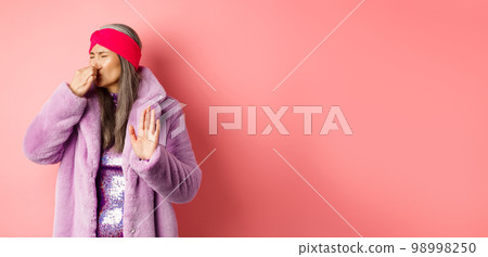 Disgusted asian mature woman in trendy purple winter coat and dress, shut nose and showing stop, refusal gesture, smell something disgusting, standing over pink background 98998250