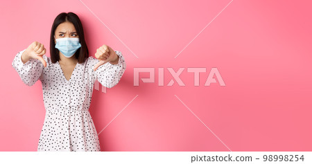 Coronavirus, social distancing and lifestyle concept. Disappointed asian woman in face mask looking judgemental, showing thumbs-down, express negative opinion, pink background 98998254