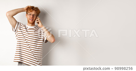 Puzzled redhead guy talking on phone, scratching head and looking confused or indecisive, standing over white background Puzzled redhead guy talking on phone, scratching head and looking confused or indecisive, standing over white background 98998256