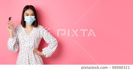 Covid-19, pandemic and lifestyle concept. Beautiful korean woman in dress and medical mask showing hand sanitizer, recommending antiseptic, standing over pink background Covid-19, pandemic and lifestyle concept. Beautiful korean woman in dress and medical mask showing hand sanitizer, recommending antiseptic, standing over pink background 98998321