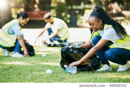 Volunteer group, nature park and cleaning for community service for clean environment, recycling and pollution. Black woman happy to help people with plastic bottle and trash bag outdoor green grass 98998542