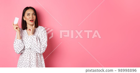 Shopping concept. Attractive asian woman thinking about buying presents, holding credit card and pondering, standing in dress against pink background Shopping concept. Attractive asian woman thinking about buying presents, holding credit card and pondering, standing in dress against pink background 98998896