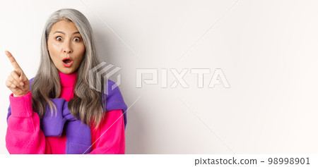 Fashion. Close up of amazed asian senior woman pointing finger upper left corner, gasping and looking impressed at camera, standing over white background Fashion. Close up of amazed asian senior woman pointing finger upper left corner, gasping and looking impressed at camera, standing over white background 98998901