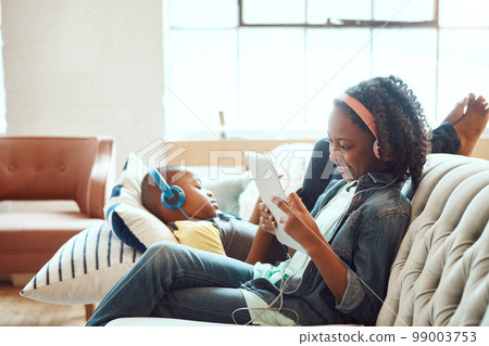 Siblings, tablet and headphones for relax on sofa in the living room enjoying entertainment or wifi at home. Happy sister and brother relaxing on couch listening to music or online streaming indoors 99003753