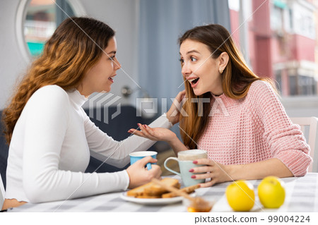 Smiling woman with cup of tea talking with girlfriend at home 99004224