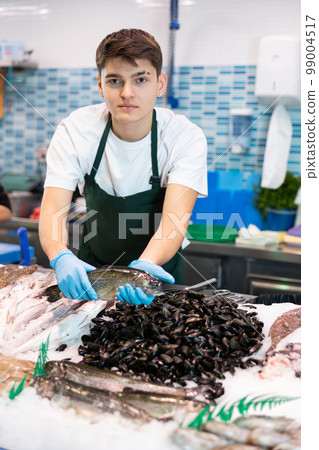Responsive shop assistant in green apron demonstrating trout in fish store 99004517