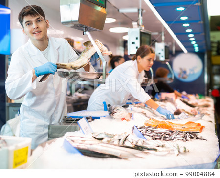 Skilful shop assistant weighing cod fish on scales in fish store Skilful shop assistant weighing cod fish on scales in fish store 99004884