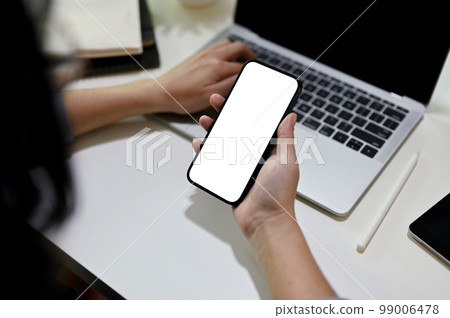 Close-up image of a female offie worker using her smartphone while using laptop at her desk Close-up image of a female offie worker using her smartphone while using laptop at her desk 99006478