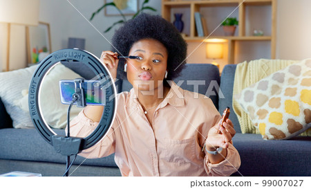 Afro beauty influencer, vlogger or podcast host talking, using phone to film live stream makeup tutorial with new mascara makeup. Excited woman using technology to promote cosmetic product from home 99007027