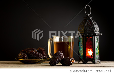 Ramadan food and drinks concept.  Ramadan Lantern with arabian lamp, wood rosary, tea, dates fruit and lighting on a wooden table with dark background. 99012551