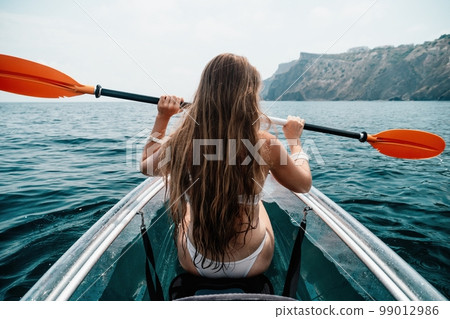 Woman in kayak back view. Happy young woman with long hair floating in transparent kayak on the crystal clear sea. Summer holiday vacation and cheerful female people relaxing having fun on the boat Woman in kayak back view. Happy young woman with long hair floating in transparent kayak on the crystal clear sea. Summer holiday vacation and cheerful female people relaxing having fun on the boat 99012986