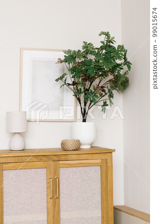 An artificial flower in a pot on a wooden chest of drawers in the living room, decorative items and a frame on the wall in a Scandinavian minimalist style 99015674