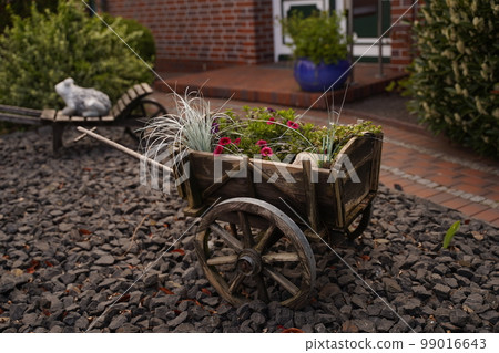 A decorative wheelbarrow stands on the stones in the garden, flowers and grass are planted in the cart. In the background is a garden path, a garden and a porch of the house. Traditional German decor. 99016643