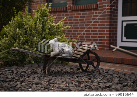 A clay frog sits in a wooden decorative wheelbarrow. In the background is a large bush, a garden and a brick house. A wheelbarrow stands in the garden. Decorative decoration for the garden. Original g 99016644