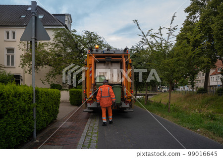 An orange garbage truck stands on a narrow green street, surrounded by green bushes and trees, a garbage man in uniform loads green garbage cans into the car. Rear view. Selective focus. Blurred backg 99016645