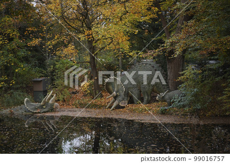 Dinosaur figures in the forest on the shore of the lake. Huge natural dinosaurs in the forest came to the watering hole. Ancient lizards in autumn forest. Dinosaur model in a natural amusement park 99016757