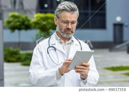 Serious mature doctor standing outside hospital, senior man in medical coat and stethoscope using tablet computer thinking and reading from online application. Serious mature doctor standing outside hospital, senior man in medical coat and stethoscope using tablet computer thinking and reading from online application. 99017358