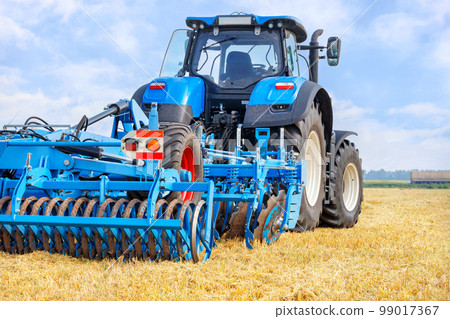 A blue agricultural tractor pulls a harrow across a mowed wheat field on a summer day. 99017367