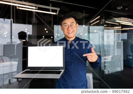 Portrait of smiling successful asian man inside office, man smiling and looking at camera standing holding laptop and pointing finger at white screen, businessman in shirt and glasses at workplace. 99017982