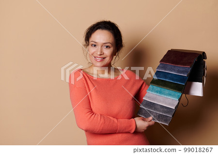 Attractive woman interior designer, smiling and showing fabric samples of upholstery materials at camera, beige backdrop 99018267