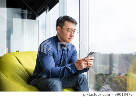 Serious thinking asian man reading news on phone, programmer using app on smartphone testing new program, businessman worker sitting on soft chair near window in modern office interior. Serious thinking asian man reading news on phone, programmer using app on smartphone testing new program, businessman worker sitting on soft chair near window in modern office interior. 99018472