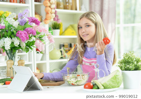 girl preparing fresh salad on kitchen table with tablet at home 99019012