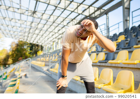 Tired hispanic athlete bent over and breathing after jogging, man doing sports on sunny day with active exercises and fitness, athlete resting after jogging. Tired hispanic athlete bent over and breathing after jogging, man doing sports on sunny day with active exercises and fitness, athlete resting after jogging. 99020541