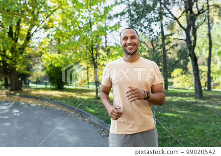 Portrait of smiling and satisfied african american man jogging in sunny park, man happy and looking at camera doing jogging and outdoor exercise. Portrait of smiling and satisfied african american man jogging in sunny park, man happy and looking at camera doing jogging and outdoor exercise. 99020542