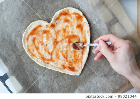 Pizza dough in the shape of a heart, the chef spreads tomato sauce on the pizza. The concept of a surprise for St. Valentine's Day. 99021044