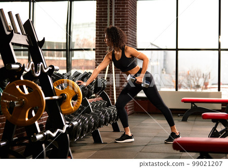 A slender athletic African American woman in sportswear does exercises with dumbbells in the gym against a mirror. The concept of sports and recreation. A slender athletic African American woman in sportswear does exercises with dumbbells in the gym against a mirror. The concept of sports and recreation. 99021207