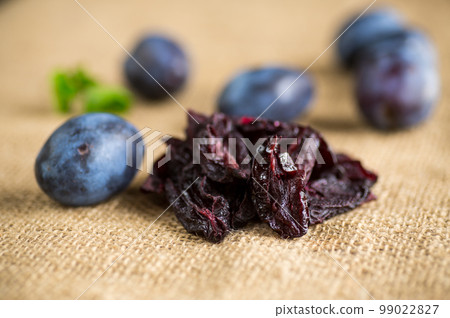 Dried prunes in a spoon on a burlap tablecloth against a background of fresh plums. 99022827