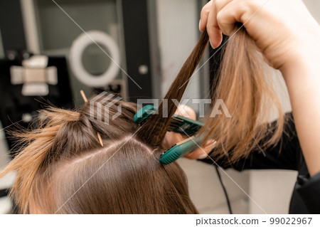 The hairdresser prepares a woman, makes a hairstyle with curls using a curling iron. Long light brown natural hair. Self care concept. The hairdresser prepares a woman, makes a hairstyle with curls using a curling iron. Long light brown natural hair. Self care concept. 99022967