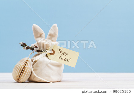 Easter bunny shaped Ivory gift bag with sweets and a gift tag with Happy Easter text and paper craft Easter egg on white wooden table, blue background banner. Willow branch 99025190