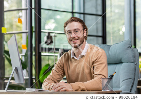 Portrait of a successful young man, programmer, IT developer, software sitting at a desk in a modern office, working at a computer. He looks at the camera, smiles. Portrait of a successful young man, programmer, IT developer, software sitting at a desk in a modern office, working at a computer. He looks at the camera, smiles. 99026184