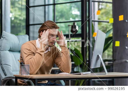 A young male programmer, IT developer codes, thinks about a new project. He is sitting concentrated at the desk in the office, holding his head thoughtfully with his hands, looking at the monitor. A young male programmer, IT developer codes, thinks about a new project. He is sitting concentrated at the desk in the office, holding his head thoughtfully with his hands, looking at the monitor. 99026195