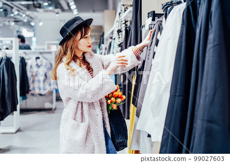 Young fashion woman customer selecting clothes. Buying clothes in boutique using fitting concept while choosing, professional stylist creating trendy look concentrated on selection in designer shop 99027603