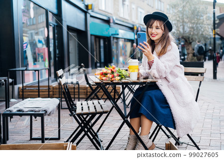 Stylish young smiling woman enjoying coffee from a reusable cup, listening to music, podcast using wireless earbuds and phone in street cafe. Fresh tulips bouquet on table. Springtime street fashion. Stylish young smiling woman enjoying coffee from a reusable cup, listening to music, podcast using wireless earbuds and phone in street cafe. Fresh tulips bouquet on table. Springtime street fashion. 99027605