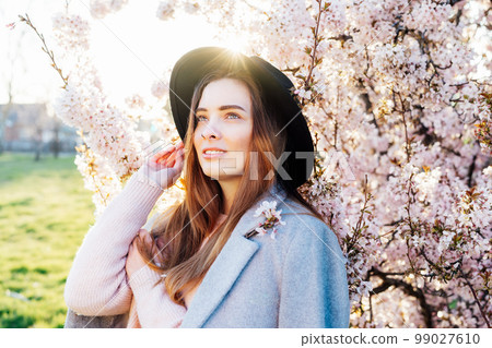 Portrait of a positive, smiling young caucasian woman in hat and coat near a blooming tree with sunset light and sunbeams. Generation Z girl enjoy springtime mood. Selective focus, copy space. Portrait of a positive, smiling young caucasian woman in hat and coat near a blooming tree with sunset light and sunbeams. Generation Z girl enjoy springtime mood. Selective focus, copy space. 99027610