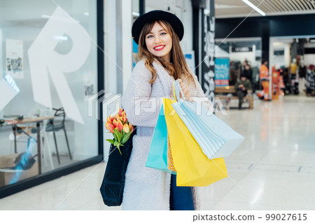 Smiling young fashion woman with net bag with fresh tulips flowers and paper shopping bags on mall background. Woman enjoying new spring season shopping. Consumerism, shopping, sale concept Smiling young fashion woman with net bag with fresh tulips flowers and paper shopping bags on mall background. Woman enjoying new spring season shopping. Consumerism, shopping, sale concept 99027615