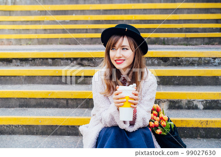 Portrait of young smiling fashion woman with fresh tulips in net bag holding reusable coffee cup while sitting on stairs at the city street. . Urban street fashion. Springtime mood. Selective focus 99027630