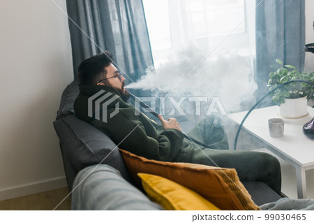 Bearded man is smoking hookah at home and blowing cloud of smoke - chill time and resting concept Bearded man is smoking hookah at home and blowing cloud of smoke - chill time and resting concept 99030465