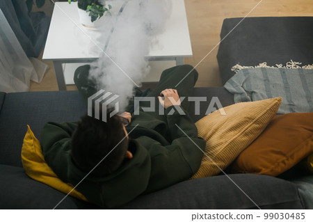 Bearded millennial or gen z man smoking hookah while relaxing on sofa at home - chill time and resting concept Bearded millennial or gen z man smoking hookah while relaxing on sofa at home - chill time and resting concept 99030485