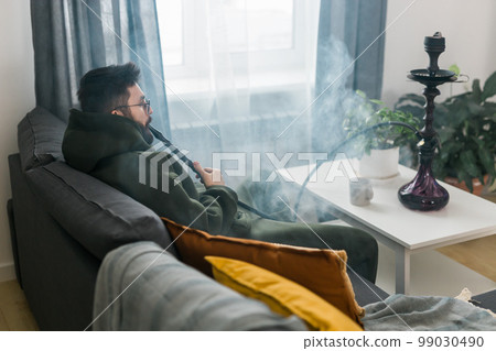Bearded millennial or gen z man smoking hookah while relaxing on sofa at home - chill time and resting concept Bearded millennial or gen z man smoking hookah while relaxing on sofa at home - chill time and resting concept 99030490