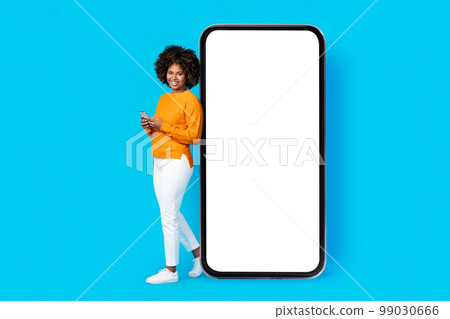 Cheerful black woman standing by huge phone white screen, mockup Cheerful black woman standing by huge phone white screen, mockup 99030666