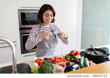 Young woman taking photo with phone while cooking in kitchen 99030733