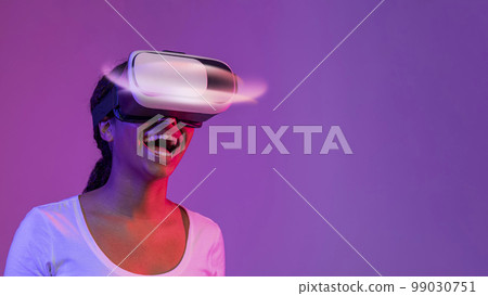 Amazed Black Lady Wearing Glowing VR Headset While Standing In Neon Light 99030751