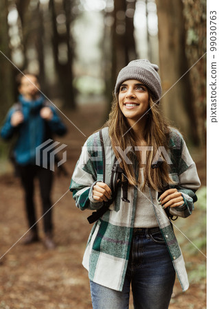 Smiling inspired young european guy and lady tourists in jackets, hat with backpack walk in autumn forest 99030763